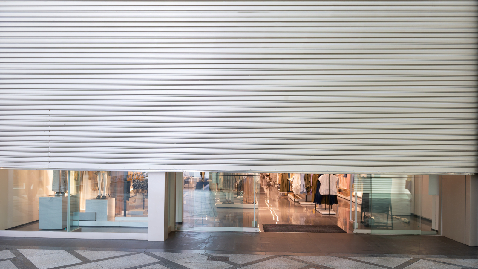A closed retail storefront featuring a metallic roller shutter, partially revealing the interior display and clothing mannequins.